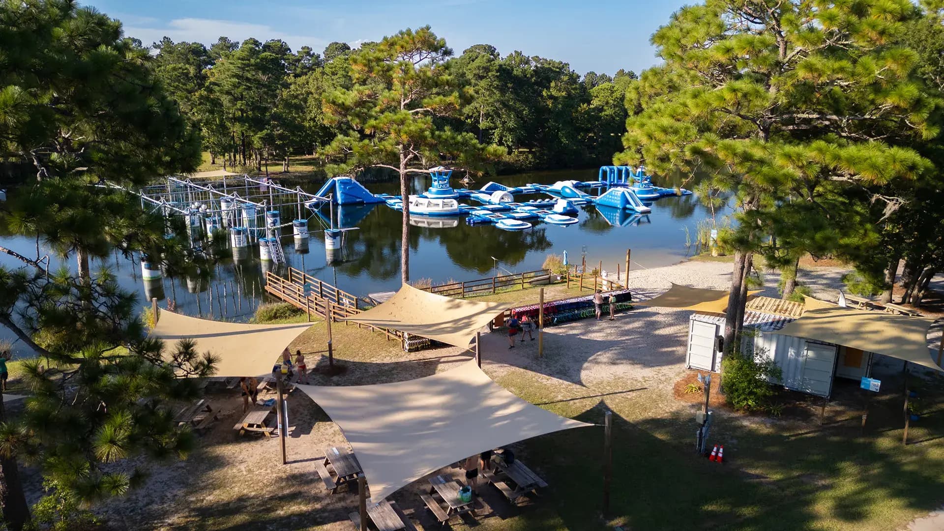 Aerial view of Charleston Aqua Park: inflatable obstacles on the lake, an aqua ropes course, and shaded picnic areas on the beach